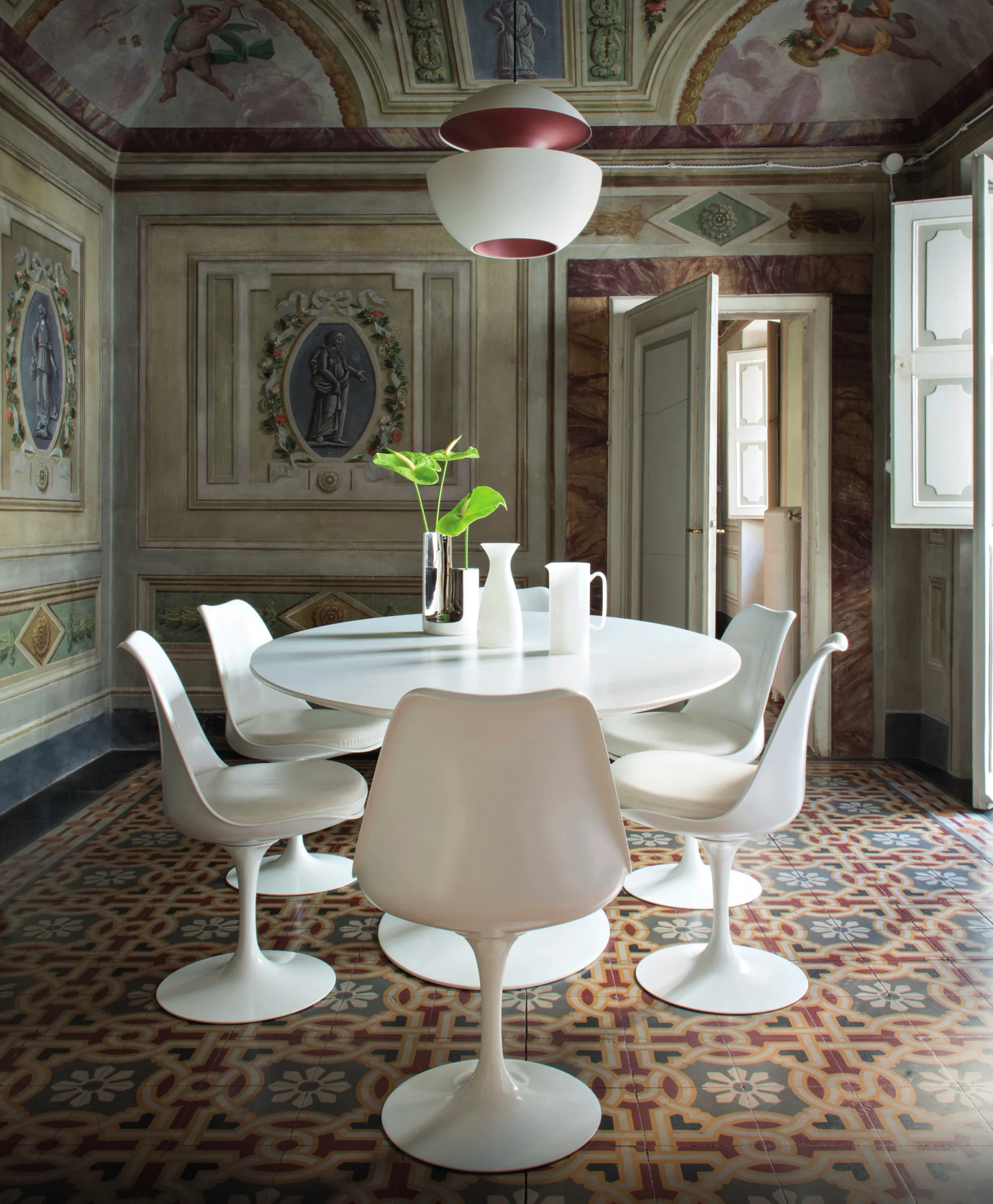 La luce dell'eleganza: sala affrescata con soffitto decorato e pavimento geometrico d'epoca, impreziosita da un tavolo rotondo e sedute bianche di design. Le finestre d'epoca originali, aperte sulla terrazza, inondano di luce l'ambiente.